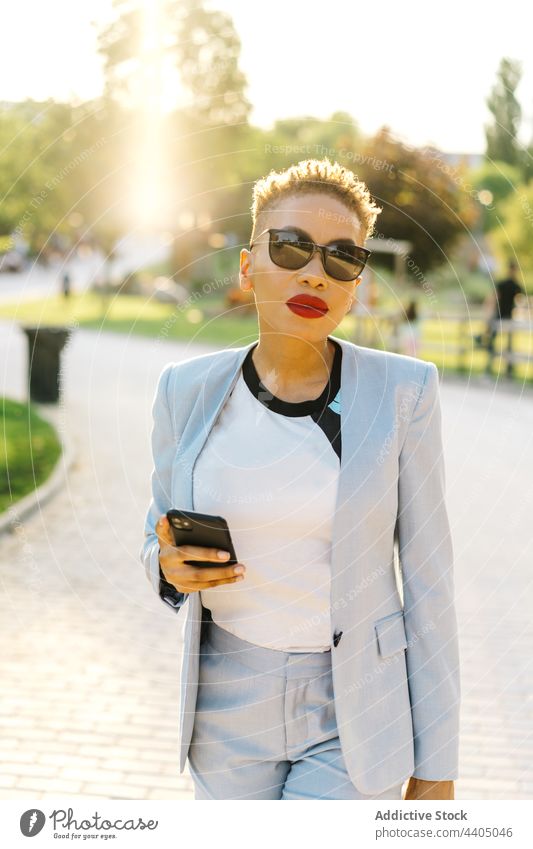 Trendy black woman chatting on smartphone while walking on pavement portrait fashion style individuality sunglasses using gadget device cellphone text messaging