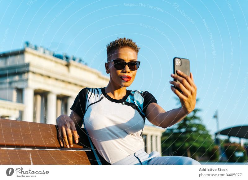 Trendy ethnic woman taking selfie on smartphone on urban bench fashion style sunglasses memory moment town using gadget device confident self portrait sit rest