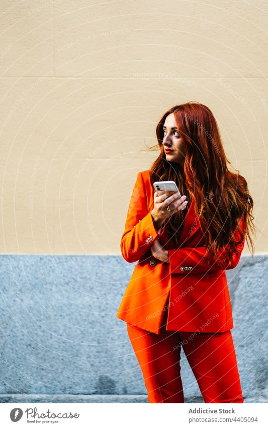 Trendy woman in orange suit browsing smartphone using color vivid redhead surfing female street style city internet outfit bright modern online contemporary