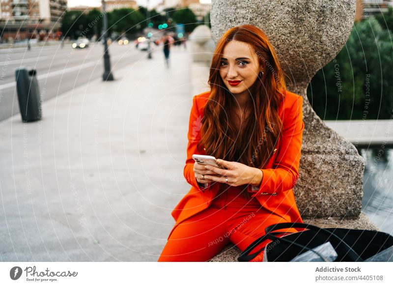 Cheerful redhead woman in orange suit browsing smartphone in street trendy color bright using female gadget style device cellphone city message modern vivid