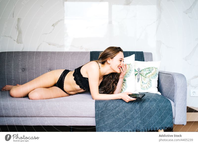 Cheerful woman in lingerie browsing smartphone in living room underwear using message cheerful sofa female home mobile social media lying slim slender content
