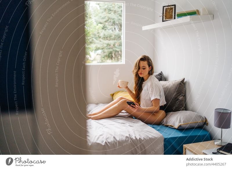 Content woman browsing smartphone on bed in morning using surfing drink cup enjoy female mobile cheerful device gadget happy internet smile coffee comfort