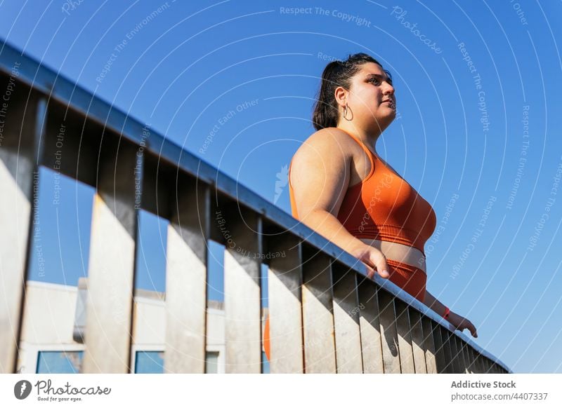 Ethnic sportswoman with excess weight behind fence in sunlight blue sky contemplative body break admire overweight pleasant alone athlete plump