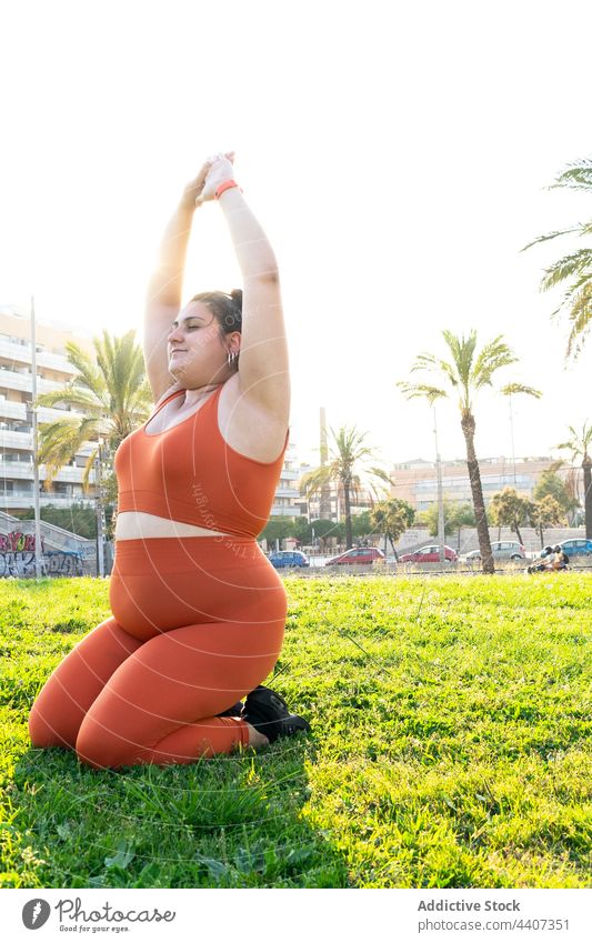 Plus size ethnic sportswoman stretching arms on lawn in sunlight athlete arms raised workout exercise training plus size warm up plump meadow sit park sky palm