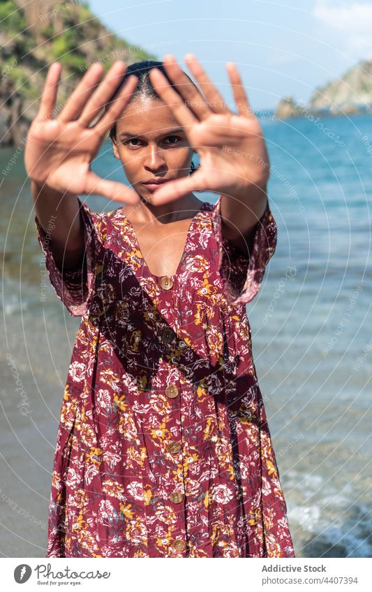 Ethnic traveler showing triangle gesture on sea shore outstretch serious sundress ornament seashore woman portrait gaze floral intent stare tourist vacation