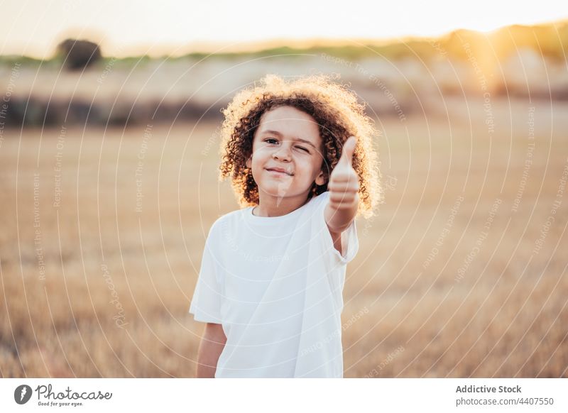 Happy ethnic kid showing thumb up in field in sunset child like gesture approve recommend wink cheerful enjoy childhood summer carefree glad happy smile content