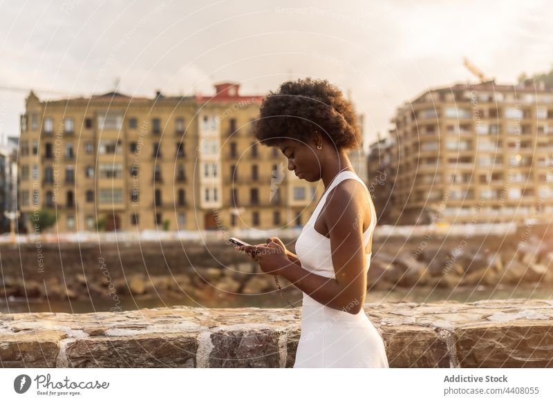 Black woman sending a message on smartphone near sea in sunset embankment using browsing cellphone summer female text black african american ethnic stand