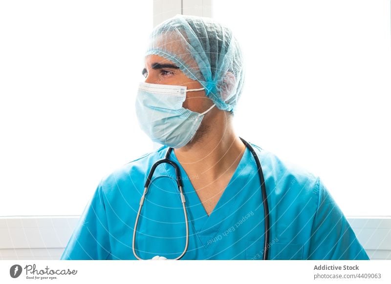 Male doctor standing in hospital with hat and face mask break medic clinic uniform tired health care man workday male job medical pause professional occupation