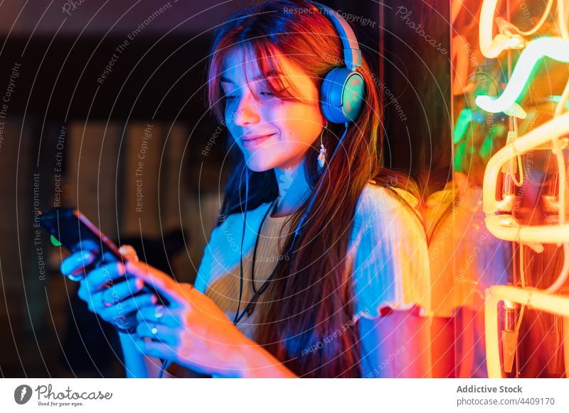 Smiling woman with smartphone listening to music from headset headphones smile spare time neon town using gadget device interested internet online watching