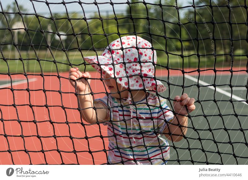 Toddler girl in a sunhat holding onto the net at a tennis court on a hot day; baby cruising and learning to walk a developmental milestone infant child