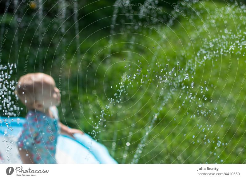 Young toddler standing in a splash pad sprinkler toy surrounded by sprays of water on a hot day; background is lush green grass pool baby pool wade vinyl