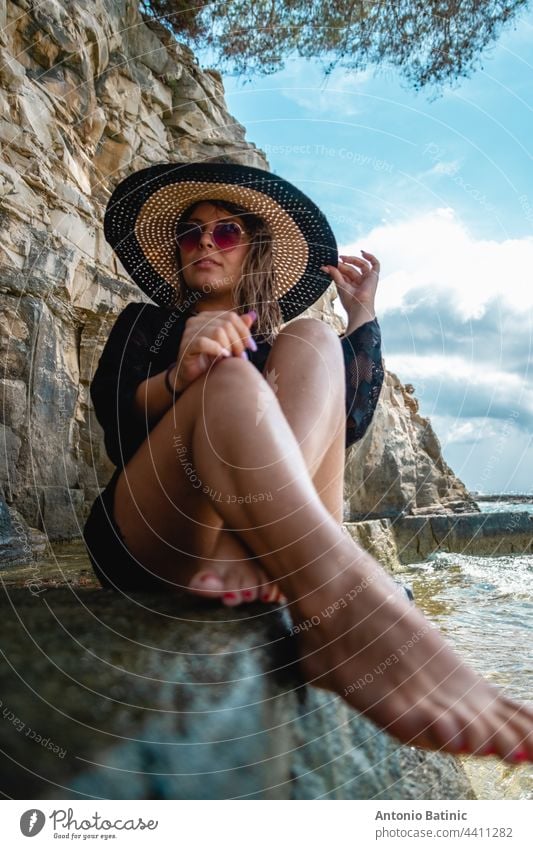 Attractive girl wearing a black dress, sunglasses and a hay hat sitting by the sea. Stormy day by the beach in Croatia, warm summer of 2021. Barefoot feet in foreground