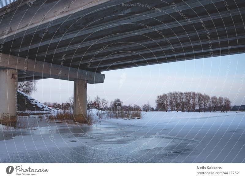 pillars under the bridge in winter on the ice of the river. big transport bridge in Latvia from under, winter evening light, clear sky, dry yellow reeds, snow covered ground