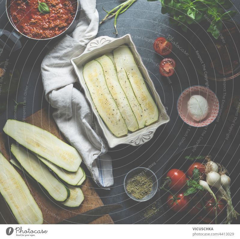 Cooking of zucchini lasagna. casserole with sliced ​​zucchini and ready-made sauce bolognese  on  kitchen table with fresh ingredients: tomatoes, mozzarella cheese and herbal salt. Top view