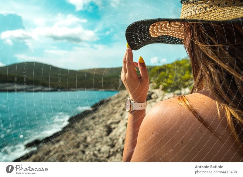 Side view of an unrecognizable brunette girl, holding onto her hay hat in the summer. Standing near the shore of the adriatic sea, concepts of travelling, summer and vacations