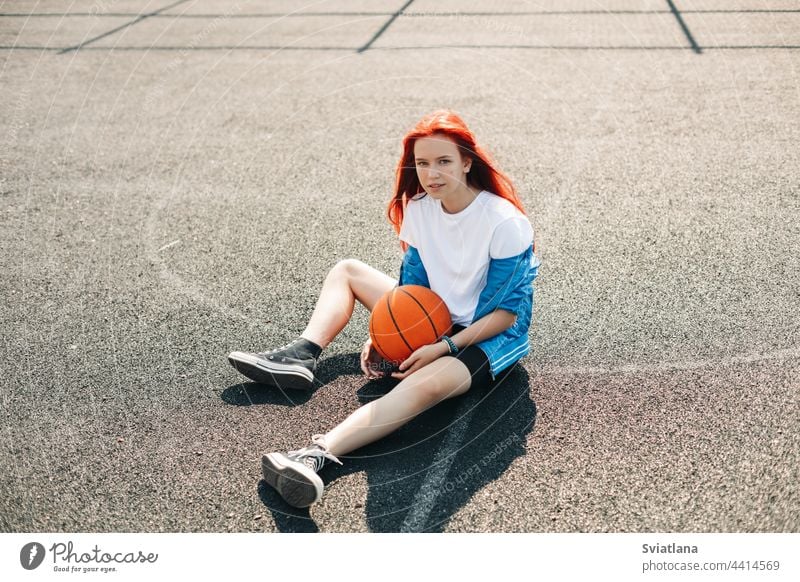A charming teenage girl with a basketball is resting after a workout. A girl on the sports field. Sports, training, healthy lifestyle sit ground playing