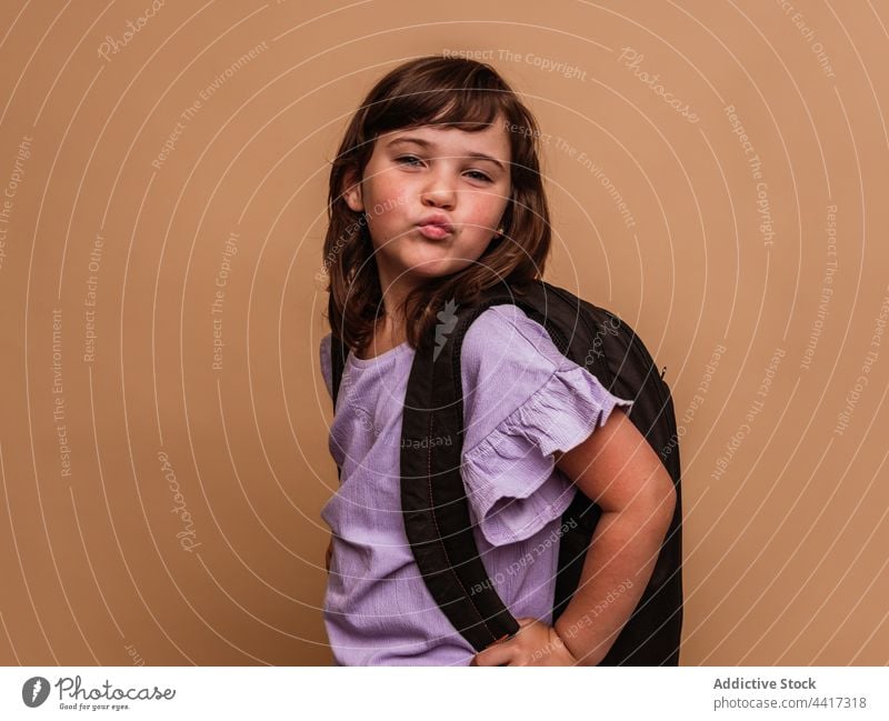 Charming girl with backpack on brown background schoolgirl pouting lips duck face child studio make face schoolchild pupil kid appearance cute adorable