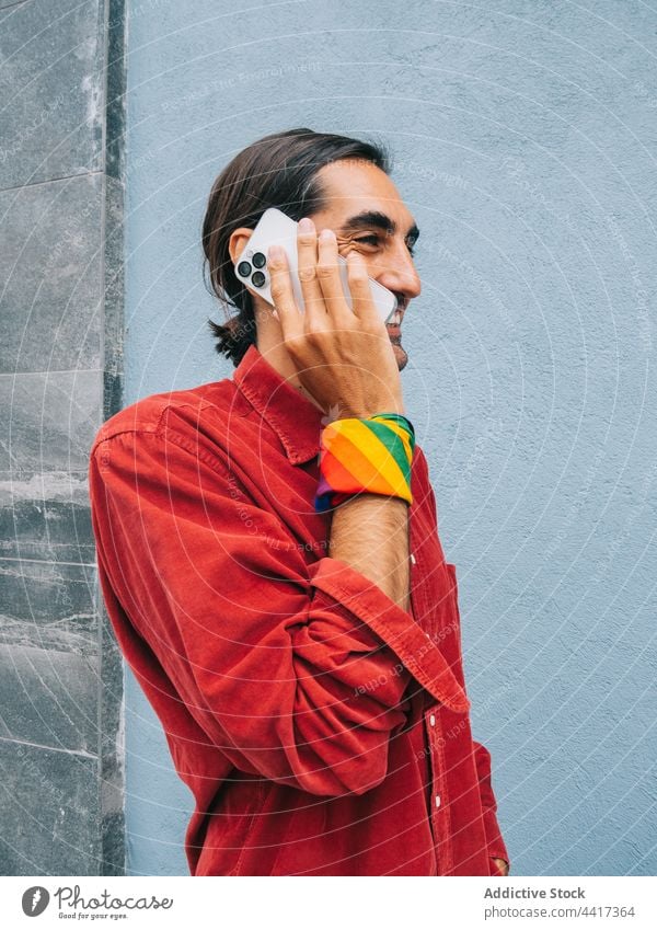 Cheerful ethnic homosexual man talking on smartphone gay city street phone call lgbt rainbow male style gadget device wall speak using smile trendy cellphone