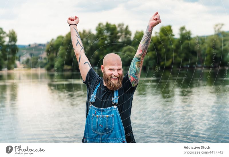Cheerful man with raised hands against lake smile nature summer hipster tattoo tree beard water fun cheerful young playful brutal happy casual male positive joy