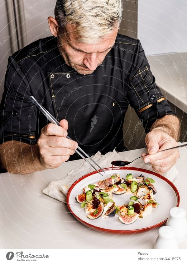 A stylish chef in a black jacket prepares tasty salad with ripe figs, goat Chevre cheese, fresh cucumber roll, beetroot and mustard leaves with tomato sauce. Restaurant dish. Delicious food