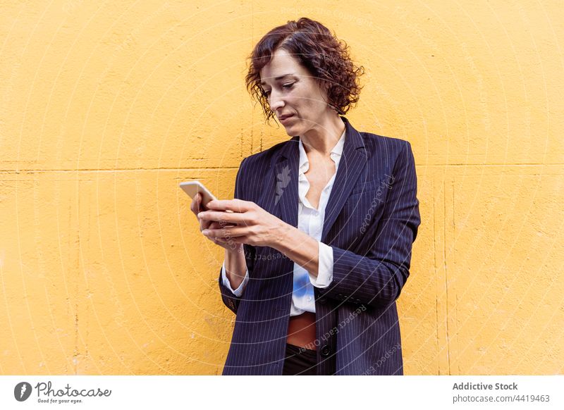 Confident well dressed woman standing browsing on smartphone near bright wall elegant formal style female classy self assured self confident happy brunette