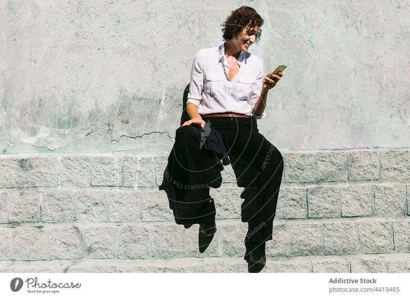 Smiling woman sitting on stone border and watching smartphone using online connection video call positive formal conversation remote female device gadget happy