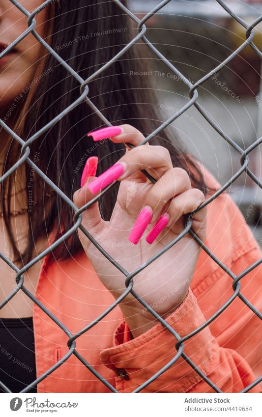 Young woman standing behind metal grid hand millennial trendy cool style manicure long nails female charming feminine brunette long hair accessory necklace