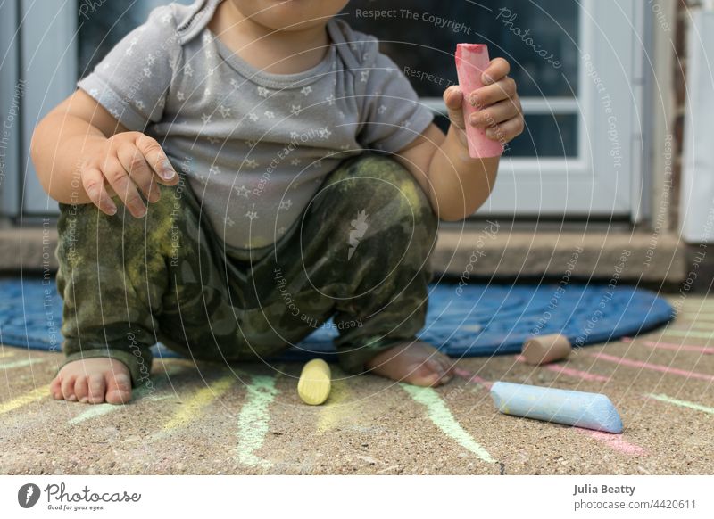 Toddler squatting while playing with chalk on the front steps of her house: child wearing camouflage print pants and star print top toddler flexible