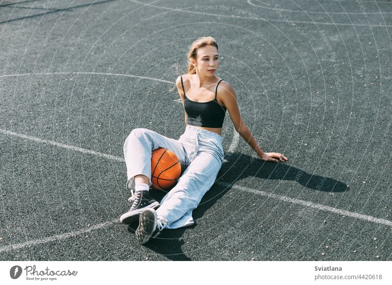 A basketball player is sitting on a sports field with a basketball and looks away. Sports, fitness, lifestyle girl playground young woman female healthy pretty