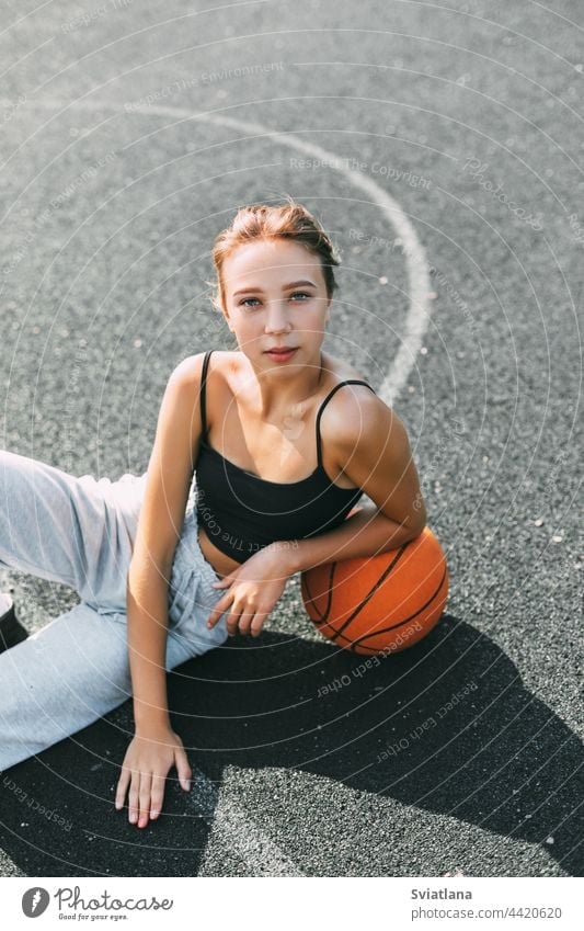 Portrait of a charming girl sitting on a sports field in a park or school with a basketball after a game or workout player playground young woman female