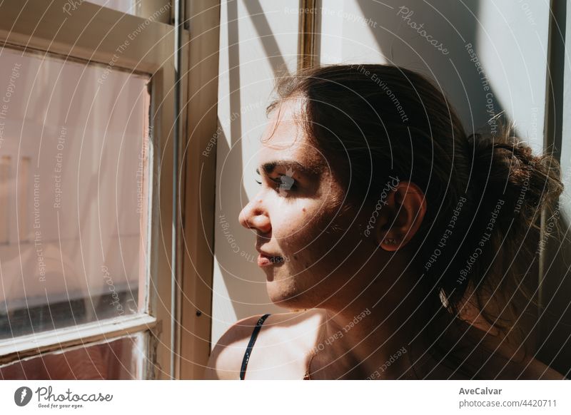 Young woman looking through a window during a super sunny day, copy space, reflexion and thinking concept, sad and anxiety, self care loneliness problems
