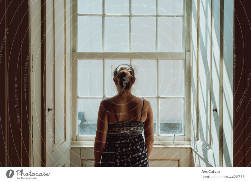Young woman using a dress looking through a window, giving the back to the camera, stress and anxiety concepts, dark image, sadness loneliness problems worried
