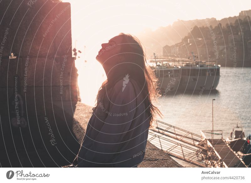Woman closing his eyes and looking up while exhaling and relaxing in the docks of the city, during a colorful sunset, mental health concept, tourism freedom