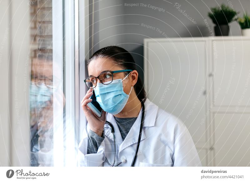 Female doctor talking on the cell phone looking out the window surgical mask serious coronavirus reflection mobile thoughtful call consulting room