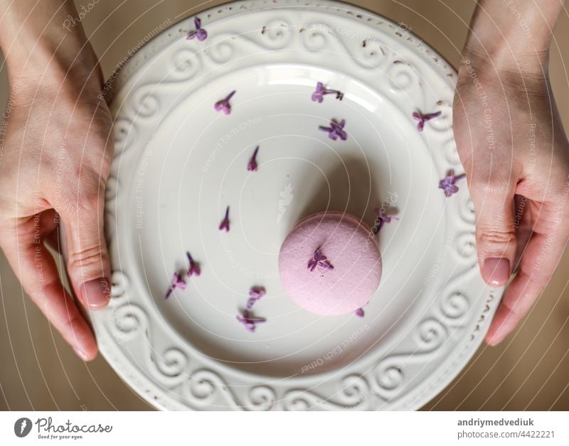 cropped view of woman holding plate with pink delicious French macaroon or macaron with lilac flowers on wooden background macarons assorted snack french bakery