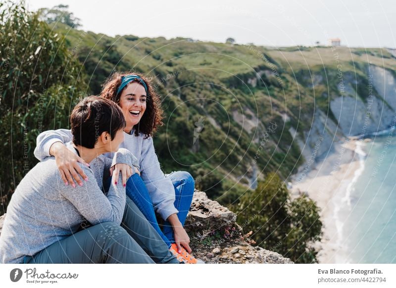 two women friends sitting, laughing and looking at beautiful sea landscape on top of the mountain. Friendship and nature concept couple cliff caucasian 30s