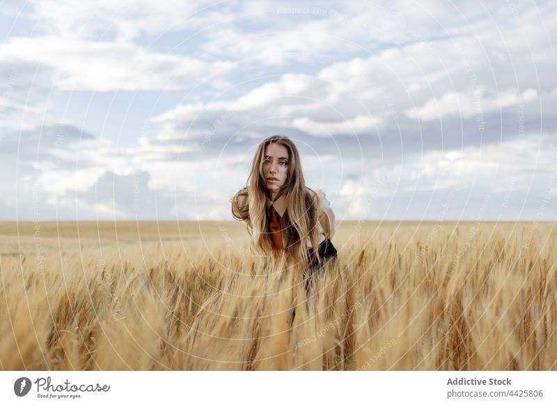 Millennial woman among wheat spikes in field nature agriculture vegetate farmland unemotional gaze portrait countryside meadow grow cereal grain cloudy sky