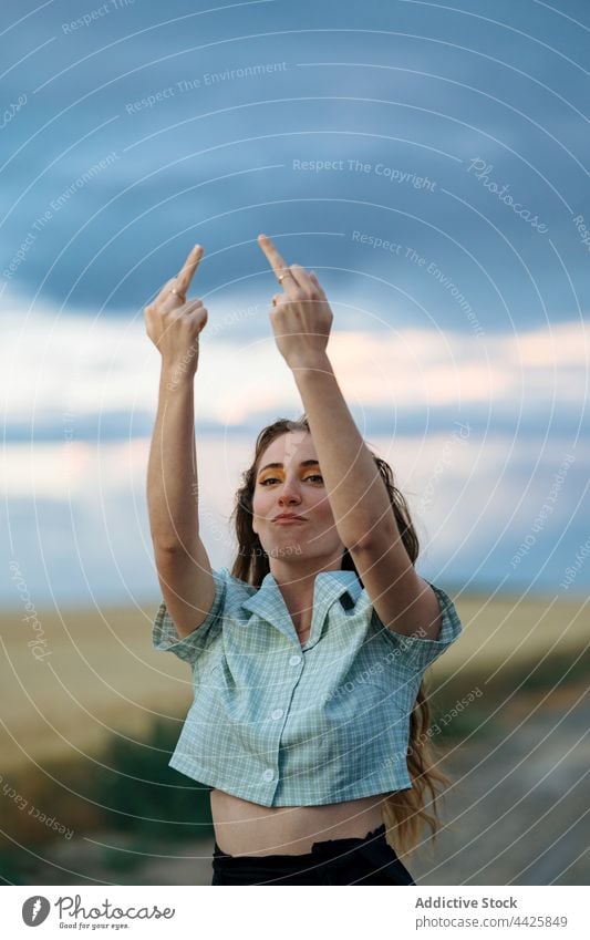 Rude woman showing fuck gesture on countryside roadway rude rebel arms raised middle finger aggressive style confident cloudy individuality evening trendy