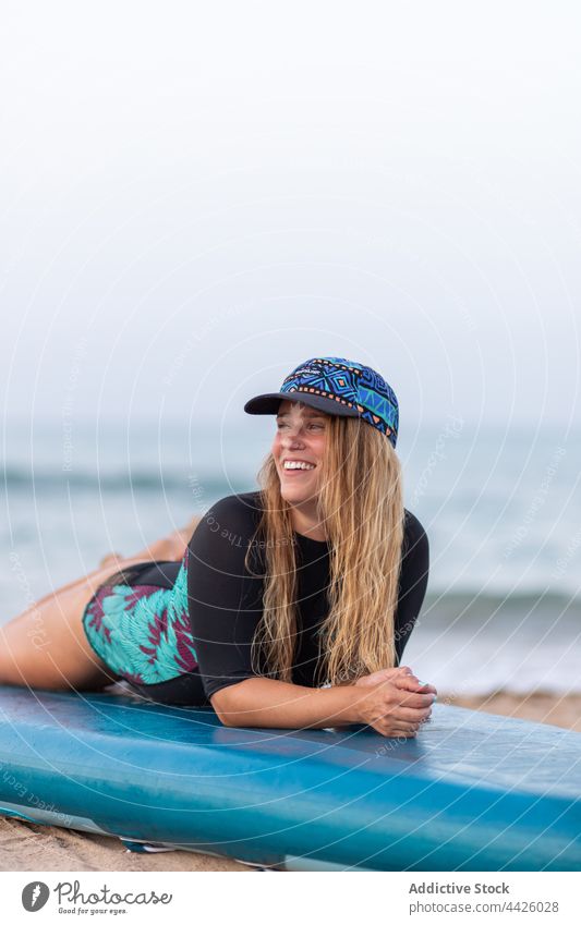 Cheerful woman lying on SUP board on beach surfer paddleboard sup board swimsuit smile seashore summer female vacation swimwear hat recreation enjoy positive