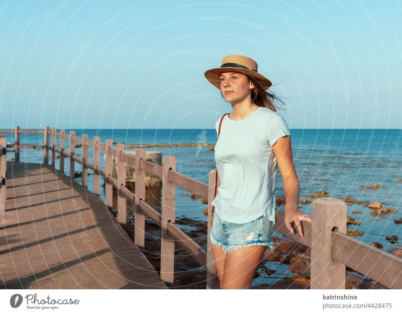 Teen girl standing on wooden bridge by the sea sunset teenager straw hat mockup Caucasian adolescent blue walkside sidewalk outdoor vacation travel look aside