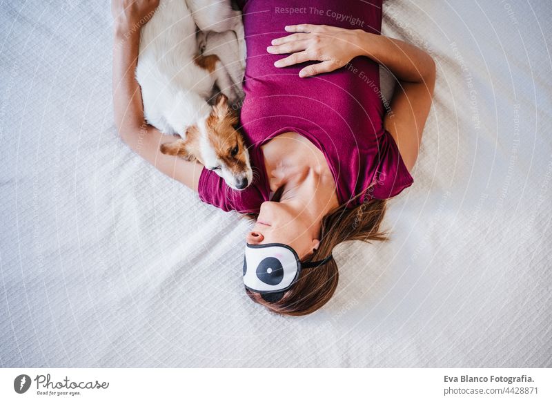 top view of relaxed caucasian woman at home wearing panda eye mask while resting on bed with cute jack russell dog. Pets, love and relax sleep kiss together