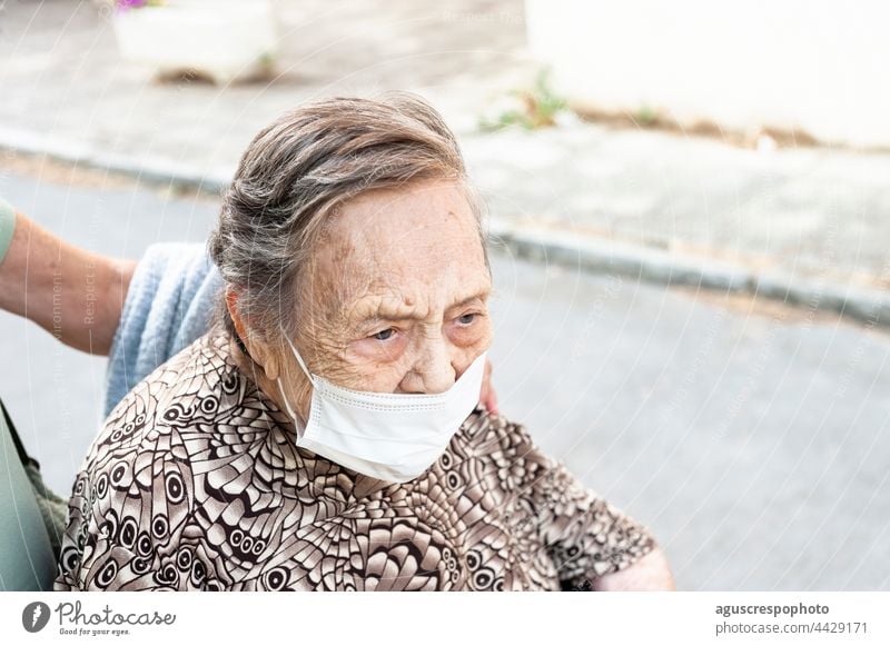 close-up of an octogenarian woman walking down the street in a wheelchair, facing forward in a patterned shirt, wearing a protective mask. grandmother face