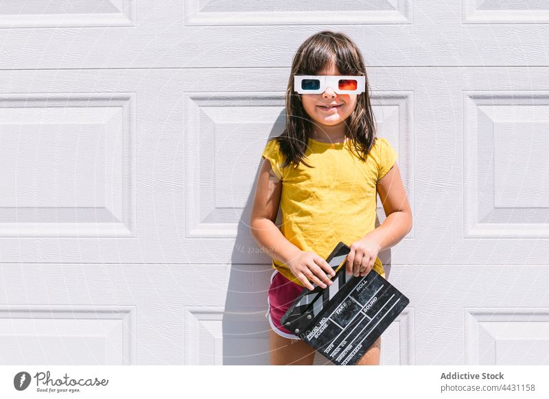 Trendy kid in 3D glasses holding clapperboard standing on wall three dimensional smile entertain 3d fun eyewear happy joy child casual girl delight watch