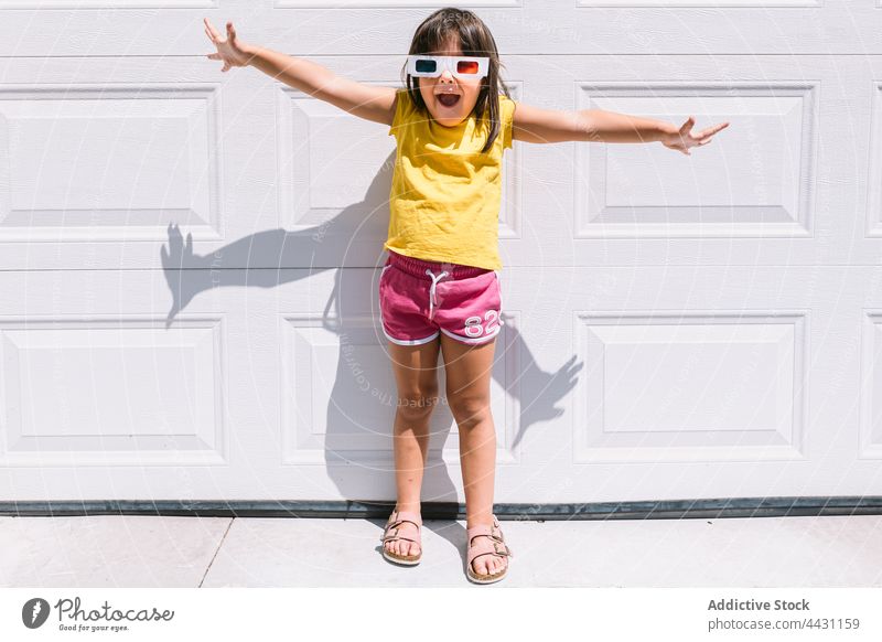 Happy trendy kid in 3D glasses standing on wall girl three dimensional entertain 3d fun happy smile eyewear showtime joy arms outstretched child casual watch