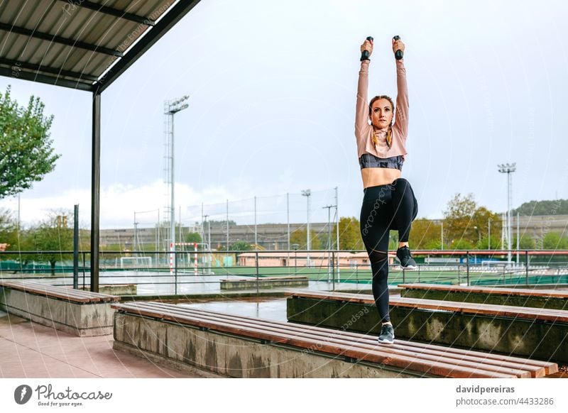 Young woman training with dumbbells outdoors arms up stretching leg knee copy space power exercise warm up young on a bench healthy female fitness girl sport