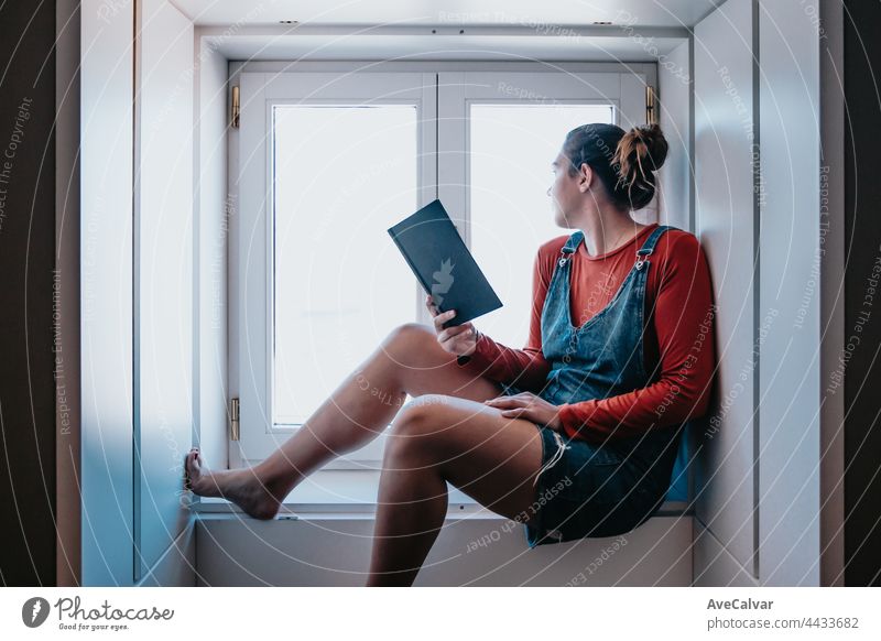 Young woman sitting next to a window reading a book during a bright day, reflexion and self care concepts, copy space resting apartment relaxation comfortable
