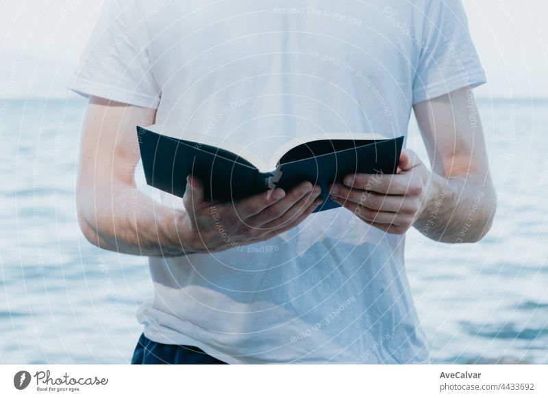 Close up of a man holding a book while reading, religious and study concepts, beach and bright day literature relaxation young adult sun sunbathing horizontal