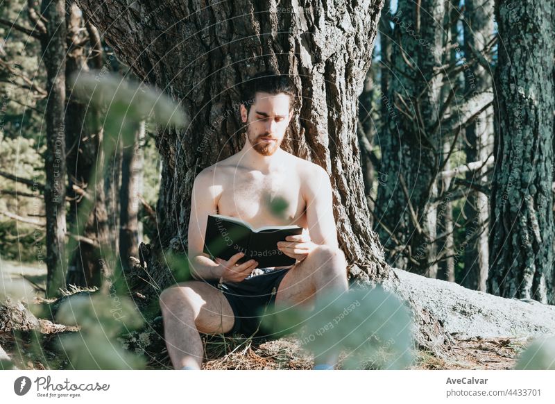 A man in trunks shirtless reading a book against a tree during a summer day, relax and chill concepts, good life, young people young adult looking at camera