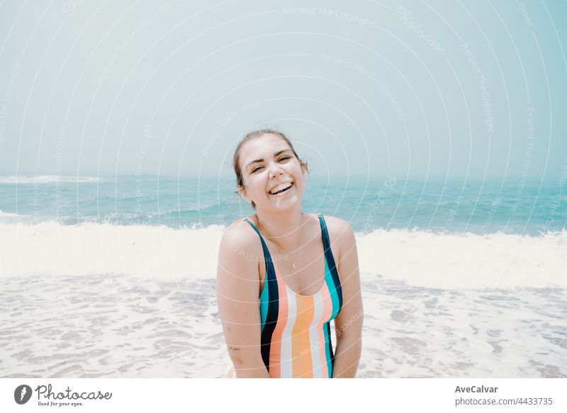 Young woman in a swimsuit laughs at camera during a sunny day at the beach with the ocean as background, liberty and holiday concept, copy space fun swimwear