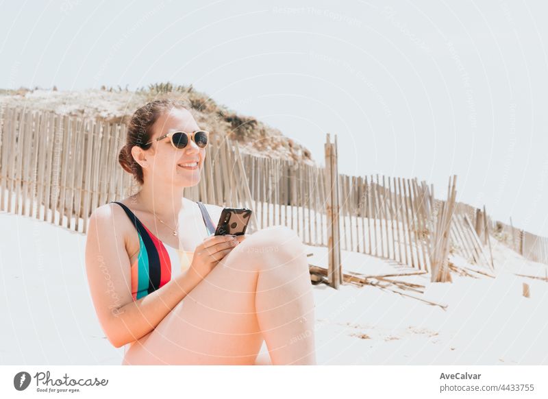 Young woman smiles while chatting on the phone wearing a colorful swimsuit at the beach taking a sun bath, travel young holiday concept, copy space, social network, sunglasses user concept